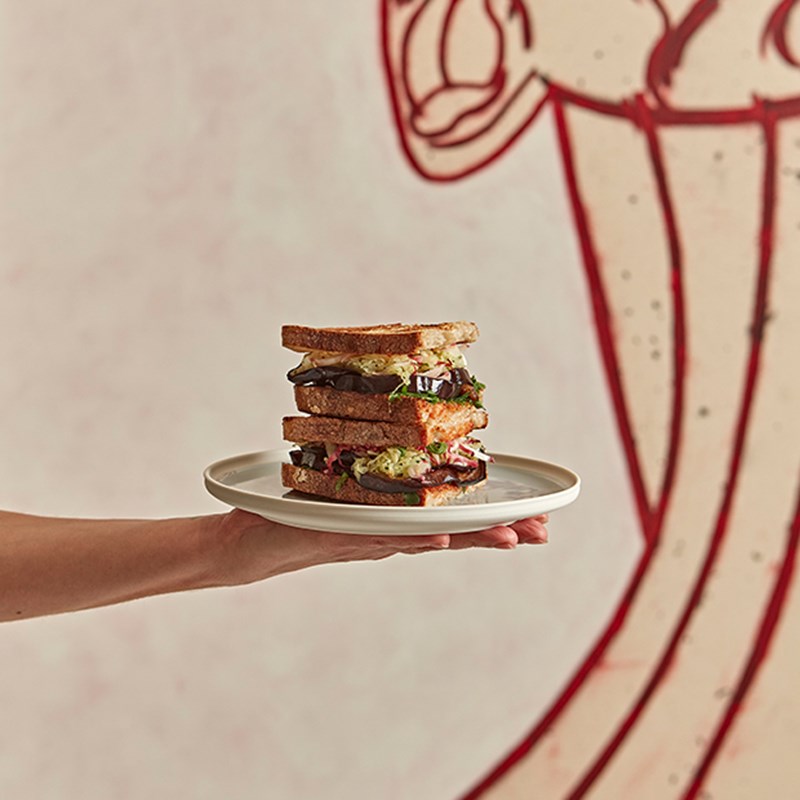 A hand holding a plate with a sandwich on it and a painting of a face in the background.