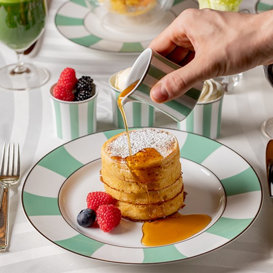Pancakes stacked on a plate with maple syrup being poured on them.